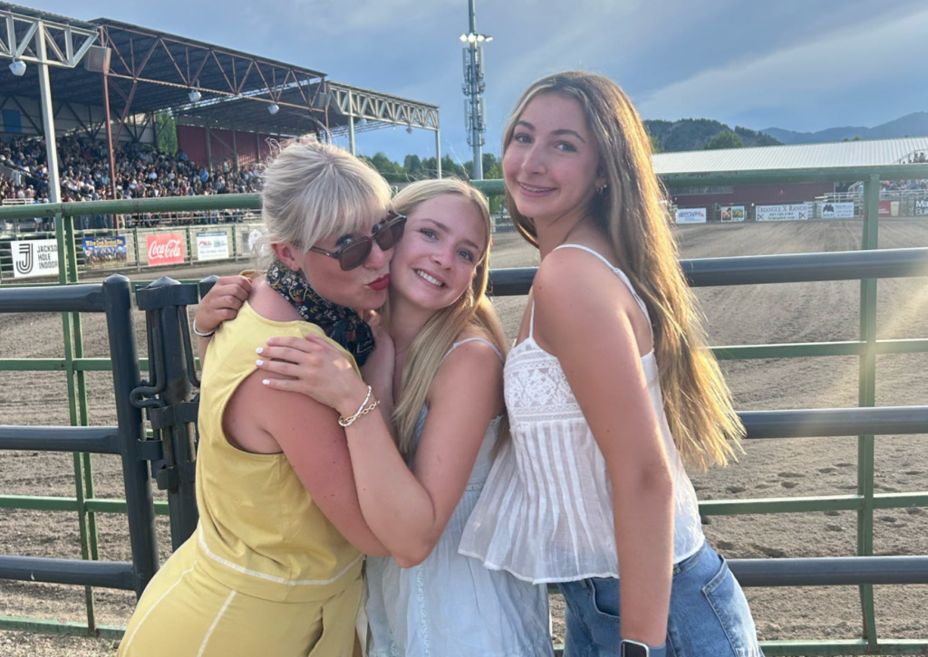 An adult and two teen girls hugging at the Jackson Hole rodeo arena railing, with the packed grandstands visible in the background