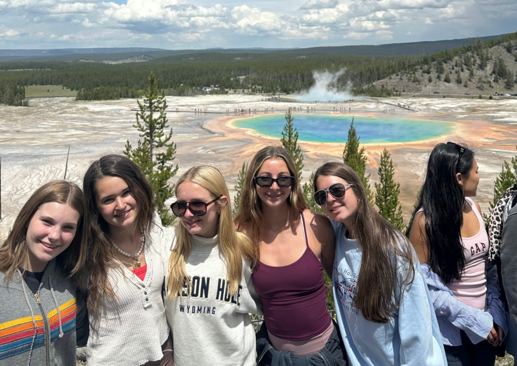 Six young women smiling and posing at the Grand Prismatic Spring overlook in Yellowstone, with the colorful rainbow-hued spring and steam visible in the background