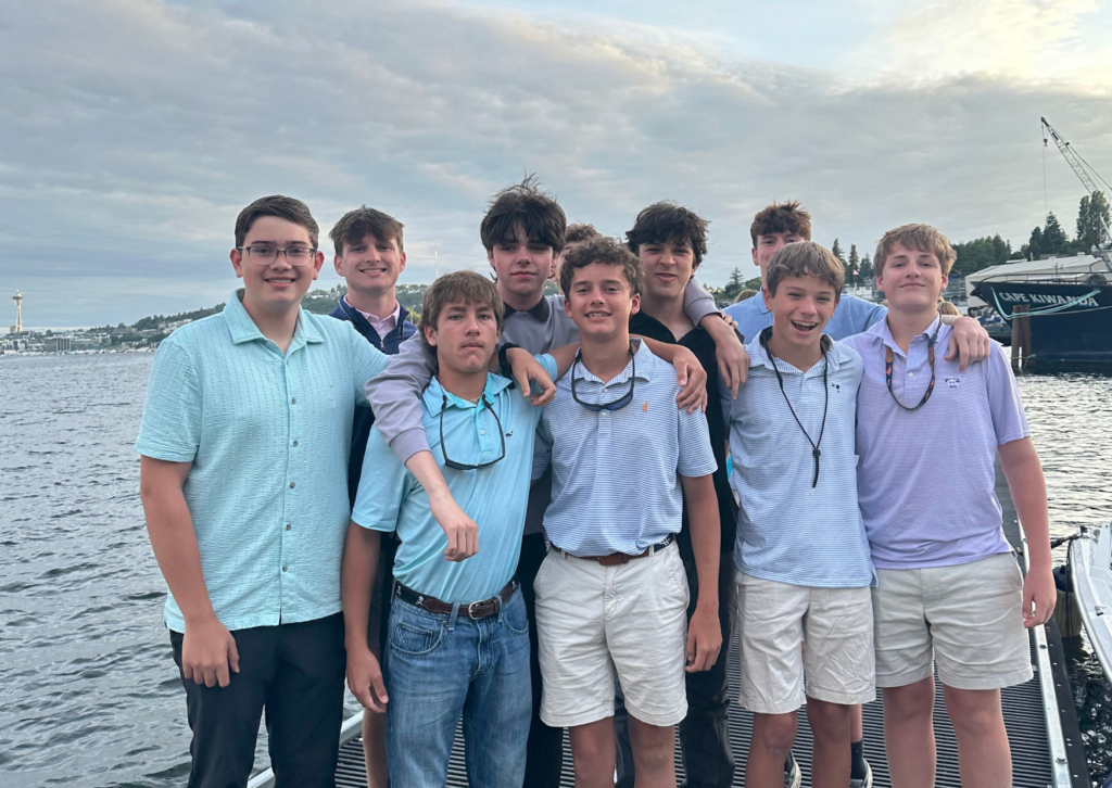 Group of male students dressed in button-down shirts posing together on a dock by the Seattle waterfront at dusk, with the Space Needle visible in the background