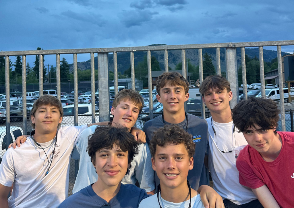 Group of seven teen boys posing together outside the Jackson Hole rodeo arena at dusk, with mountains visible in the background