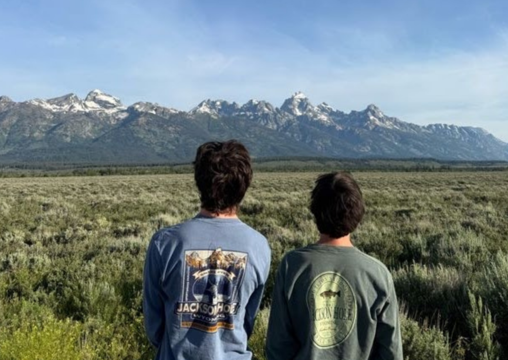 Two tour participants viewed from behind, looking out at the snow-capped Grand Teton mountain range across a sagebrush meadow at dusk