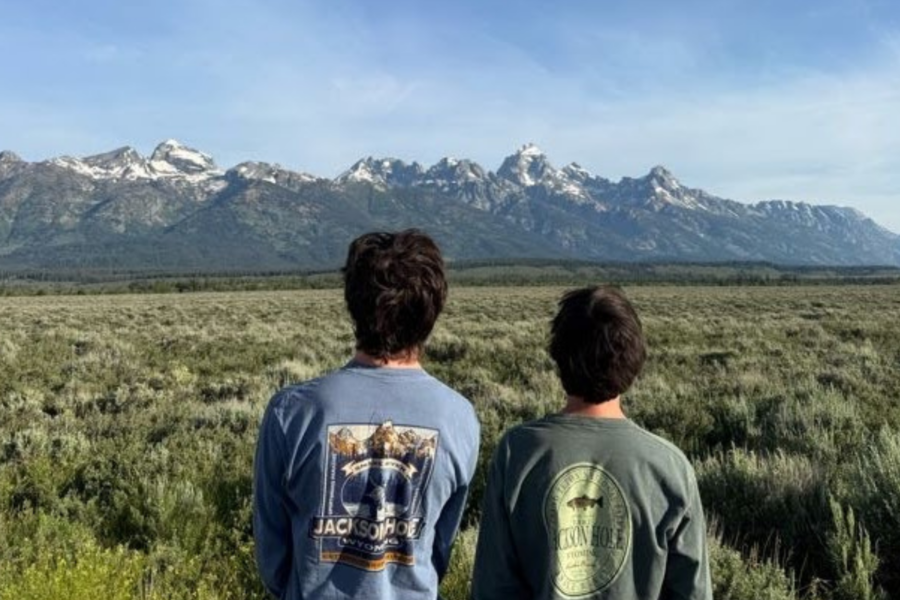 Two tour participants viewed from behind, looking out at the snow-capped Grand Teton mountain range across a vast sagebrush meadow
