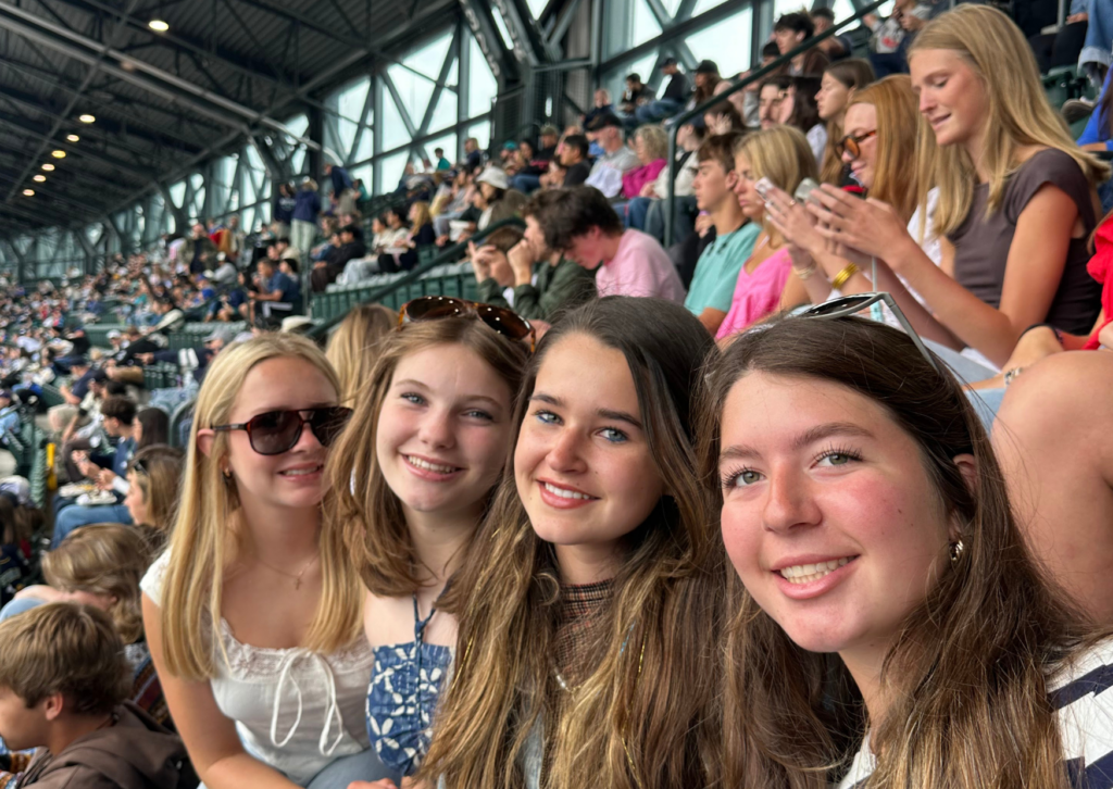 Four students smiling together in the stands at a Seattle Mariners game, with a full stadium visible behind them