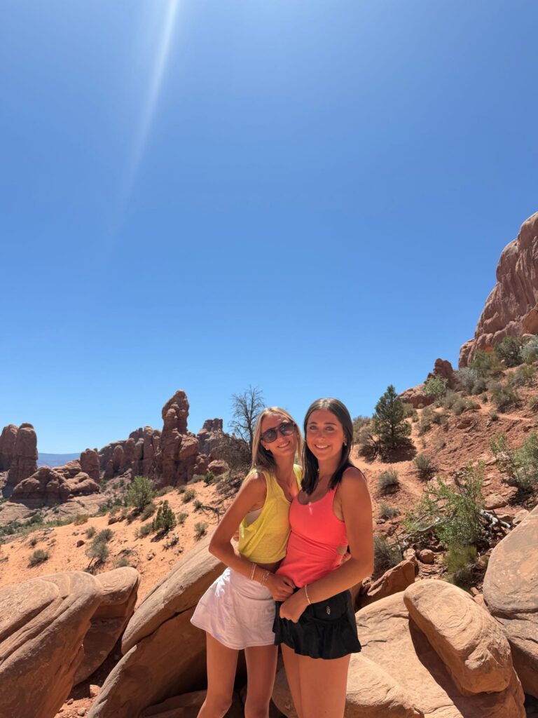 Two young women smiling and posing together on red sandstone rocks with desert rock formations in the background at Arches National Park