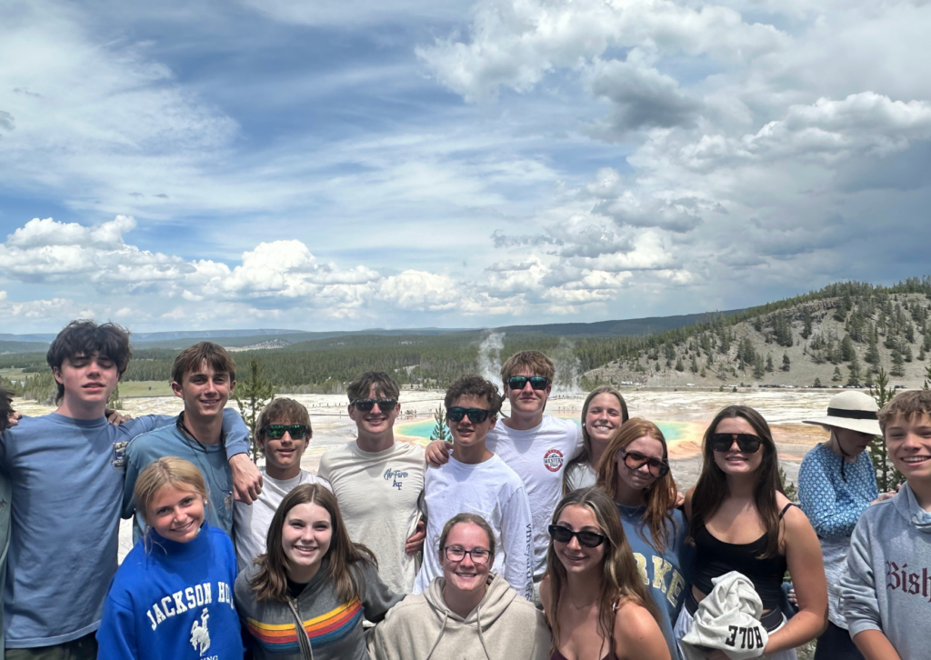 Large group of tour participants posing together at the Grand Prismatic Spring overlook in Yellowstone, with the colorful thermal spring and steam visible behind them