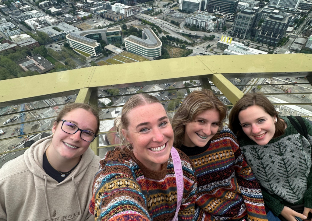 Four women taking a selfie at the Space Needle observation deck with an aerial view of Seattle below