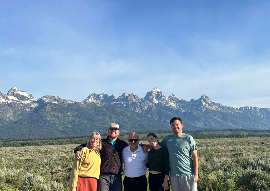 Five people smiling and posing together in a sagebrush meadow with the Grand Teton mountain range in the background