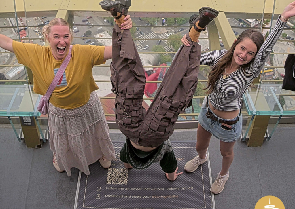Three people posing playfully on the Space Needle observation deck — one doing a handstand while two others cheer on either side, with the Seattle skyline visible below through the glass barrier