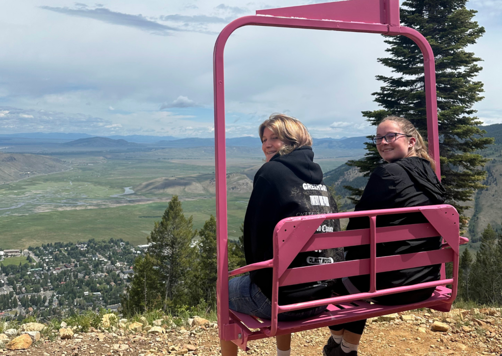 Two Grand Western Tour travelers sitting on a pink ski lift chair at the top of Snow King Mountain, with an aerial view of Jackson and the valley below