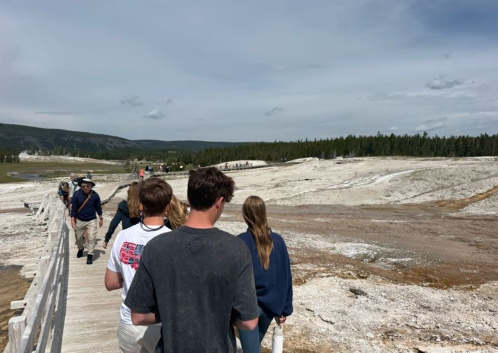Tour participants walking along a wooden boardwalk through Yellowstone's Upper Geyser Basin, surrounded by white mineral deposits and geothermal features