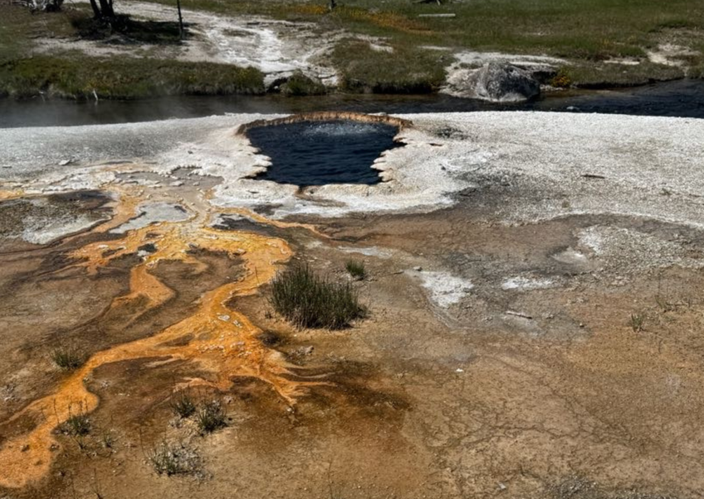 Close-up aerial view of a Yellowstone hot spring with a deep blue pool surrounded by vivid orange and yellow mineral deposits flowing across a white silica landscape