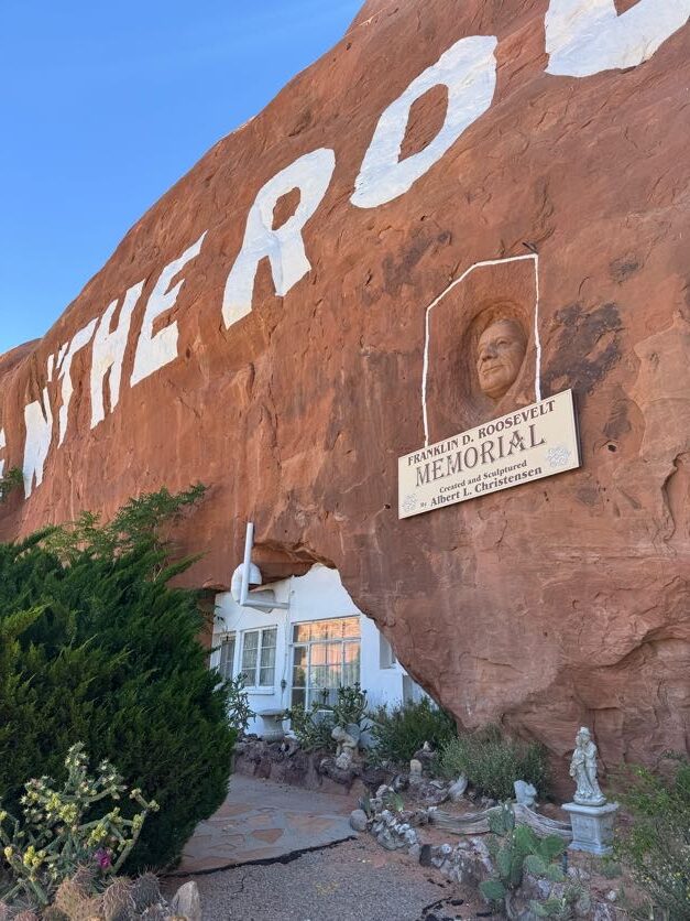 Close-up of Hole N' the Rock near Moab, showing the home carved into the cliff face with a Franklin D. Roosevelt Memorial relief carving and painted lettering on the red sandstone