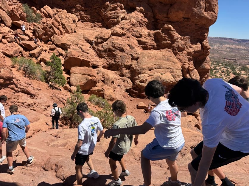Students climbing up red sandstone rocks at Arches National Park with dramatic rock formations and a desert valley visible in the background