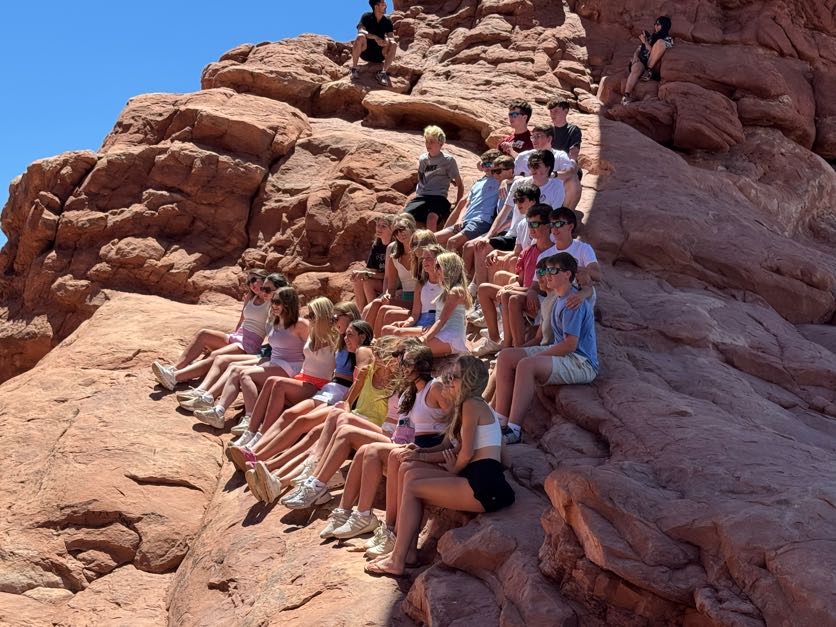 Large group of tour students sitting together for a group photo on a sloped red sandstone rock formation at Arches National Park