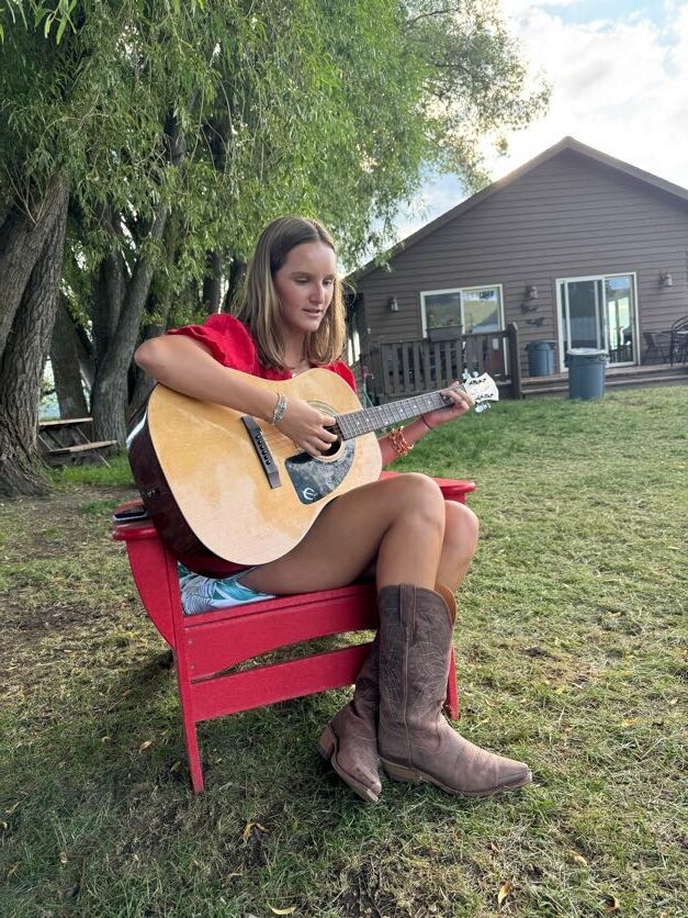 girl playing the guitar in cowboy boots outside the cabin at the ranch