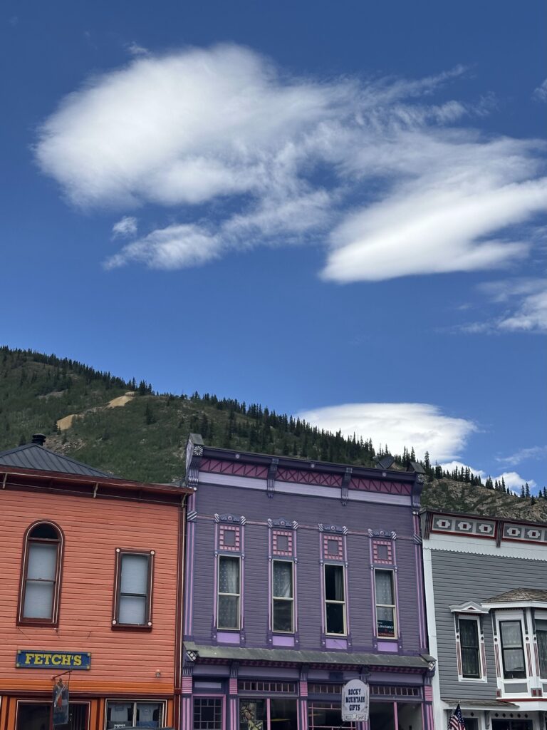 Colorful Victorian-era commercial buildings on Silverton's main street, including an orange-red and purple storefront, with a forested mountain ridge and dramatic clouds behind