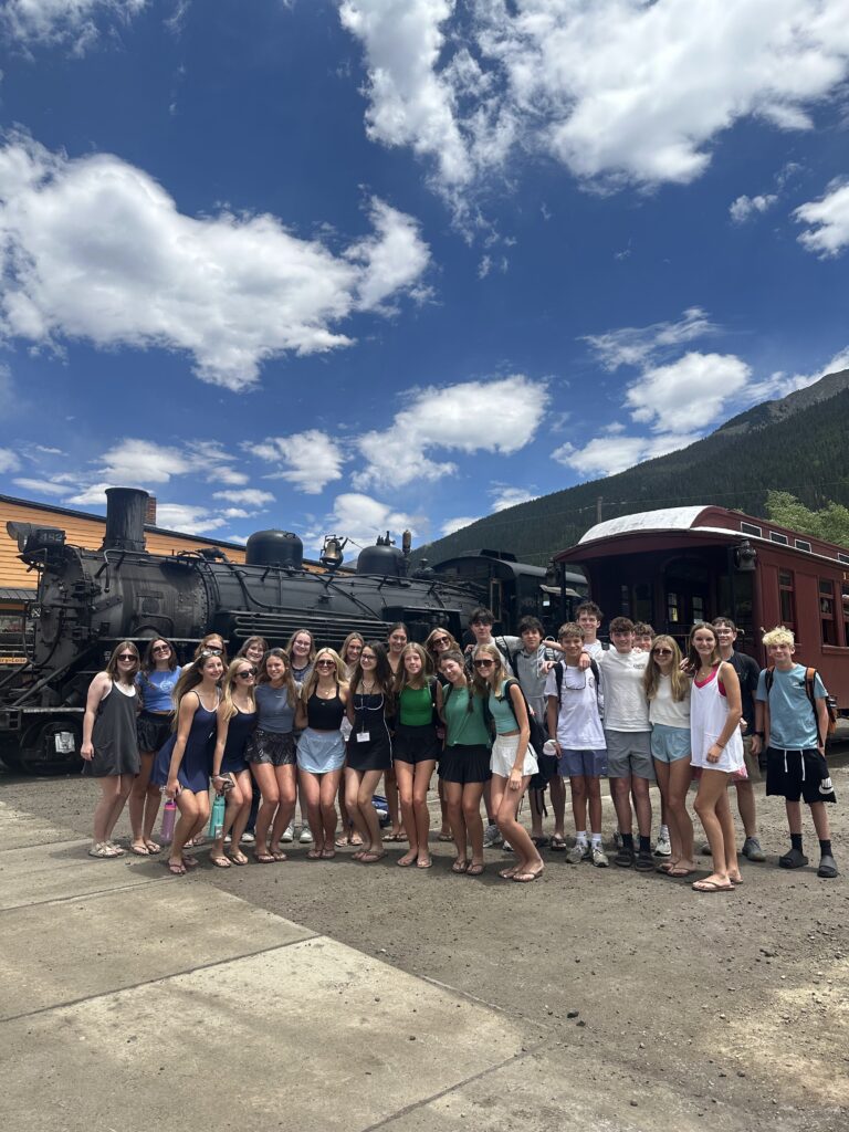 Large group of teens posing in front of a black steam locomotive and red passenger car at the Durango & Silverton Narrow Gauge Railroad depot with mountain backdrop