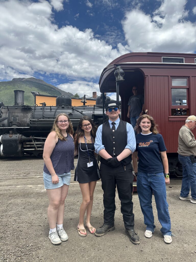 Three smiling teen girls posing with a train conductor in uniform—black vest, tie, and hat—in front of the steam locomotive at the Durango & Silverton depot