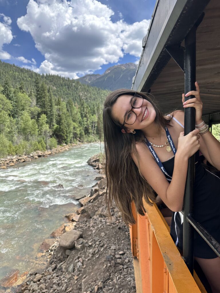 Smiling teenage girl with glasses and long hair leaning out from the open side of the train car holding the railing, with the blue-green Animas River and forested mountain slopes visible