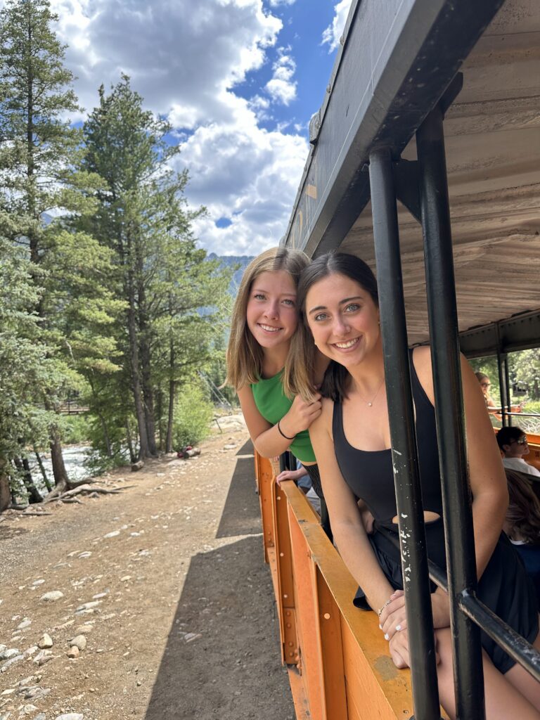 Two smiling teenage girls leaning out from the open side of the train car with pine trees and mountain terrain visible