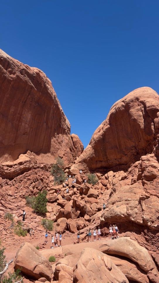Towering red sandstone rock formations at Arches National Park with small figures of hikers visible at the base, showing the massive scale of the landscape