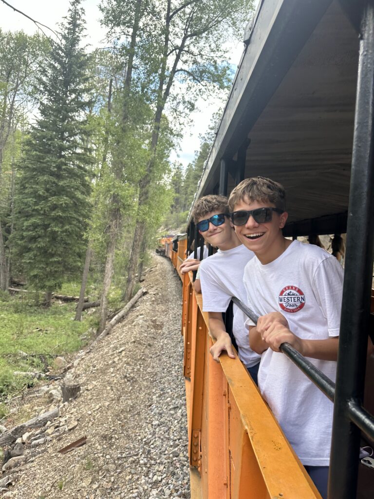 Two grinning teen boys in sunglasses leaning from the open side of the train car, one wearing a Grand Western Tour t-shirt, with pine-forested mountain slope visible