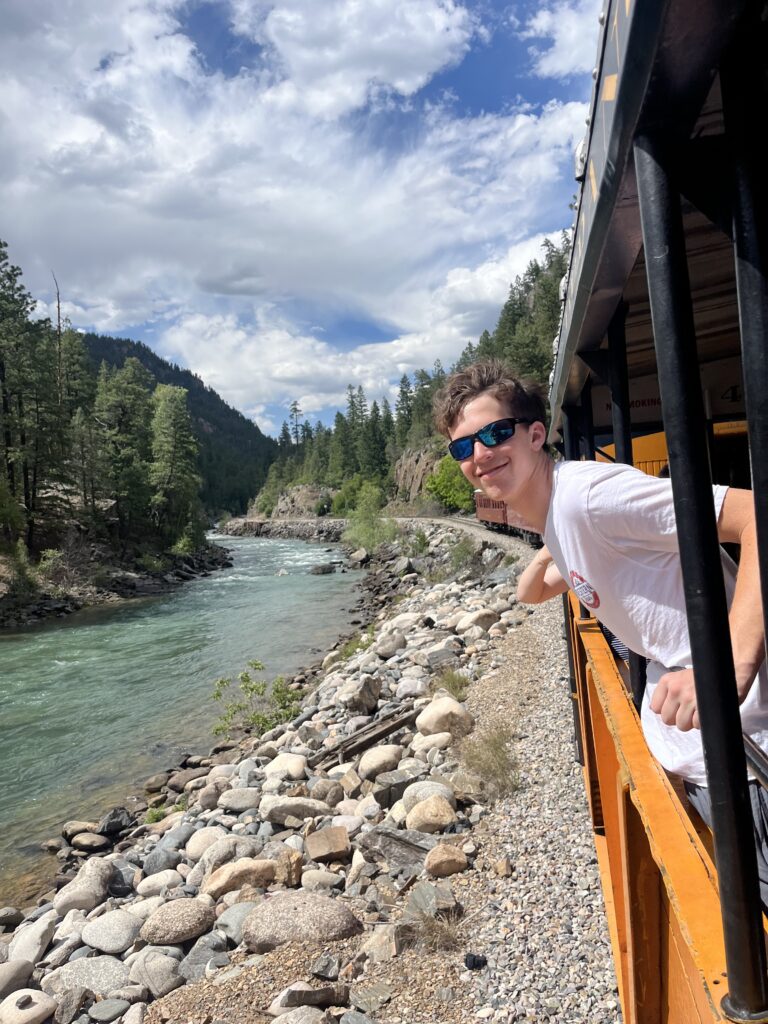 Smiling teen boy in sunglasses leaning out from the train car over the rocky bank of the Animas River with forested mountain canyon behind