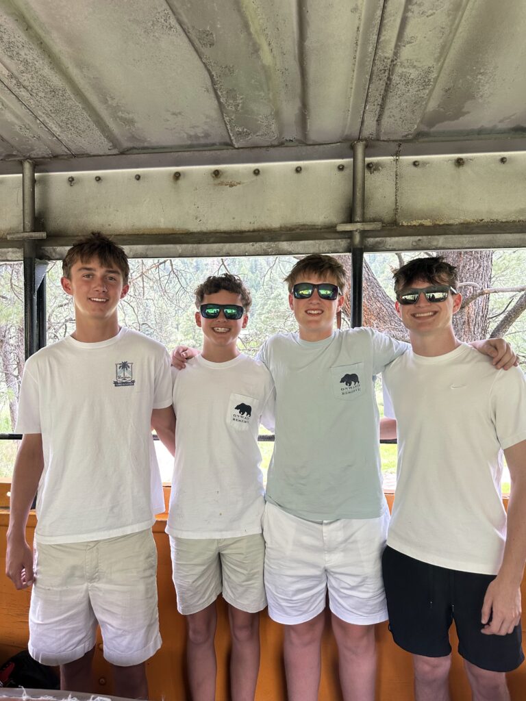 Four smiling teen boys in white t-shirts and sunglasses posing together inside the open-air train car with forested mountain scenery through the windows