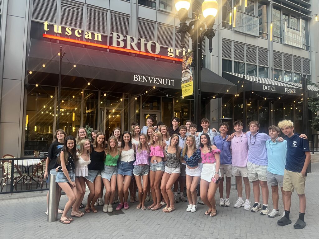 Large group of teens posing in front of Brio Grille restaurant in Durango in the evening, with string lights and Benvenuti signage above the entrance