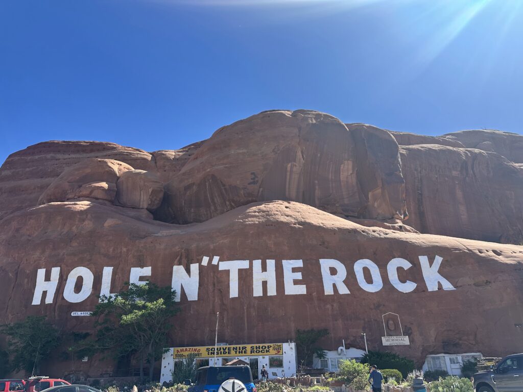 Wide exterior view of Hole N' the Rock attraction near Moab, with large white painted lettering reading HOLE N' THE ROCK on the sandstone cliff and a souvenir shop below