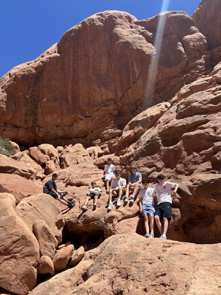 Small group of young men resting and sitting on red sandstone rocks at the base of a towering cliff face at Arches National Park