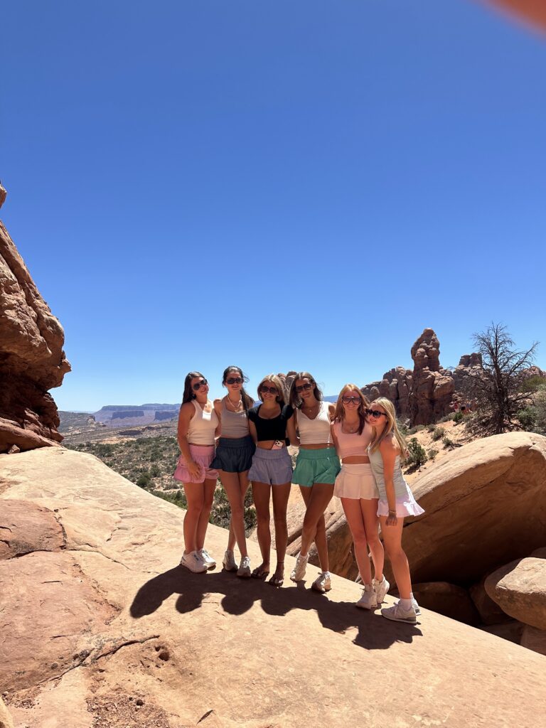 Group of five young women posing on top of a red sandstone rock at Arches National Park with a wide desert canyon view and blue sky behind them