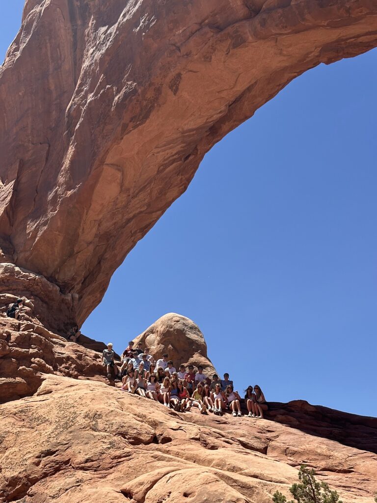 Tour group posing together on a sloped rock ledge beneath a massive natural sandstone arch at Arches National Park, Utah