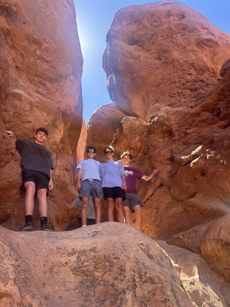 Four students posing in a narrow rocky crevice between towering red sandstone formations at Arches National Park