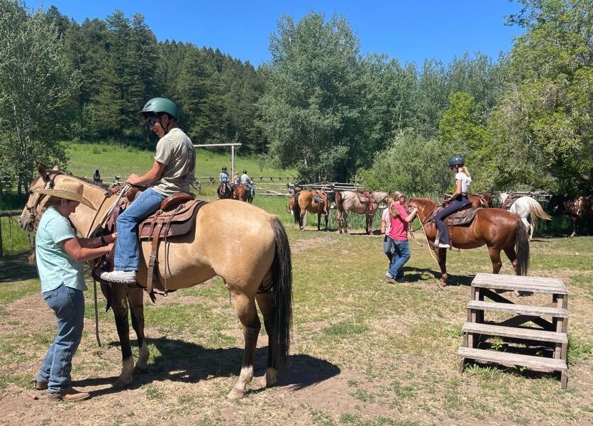 Ranch instructor adjusting a student's stirrups while the student sits on a palomino horse, with other riders and horses gathering in the background at Granite Creek Ranch