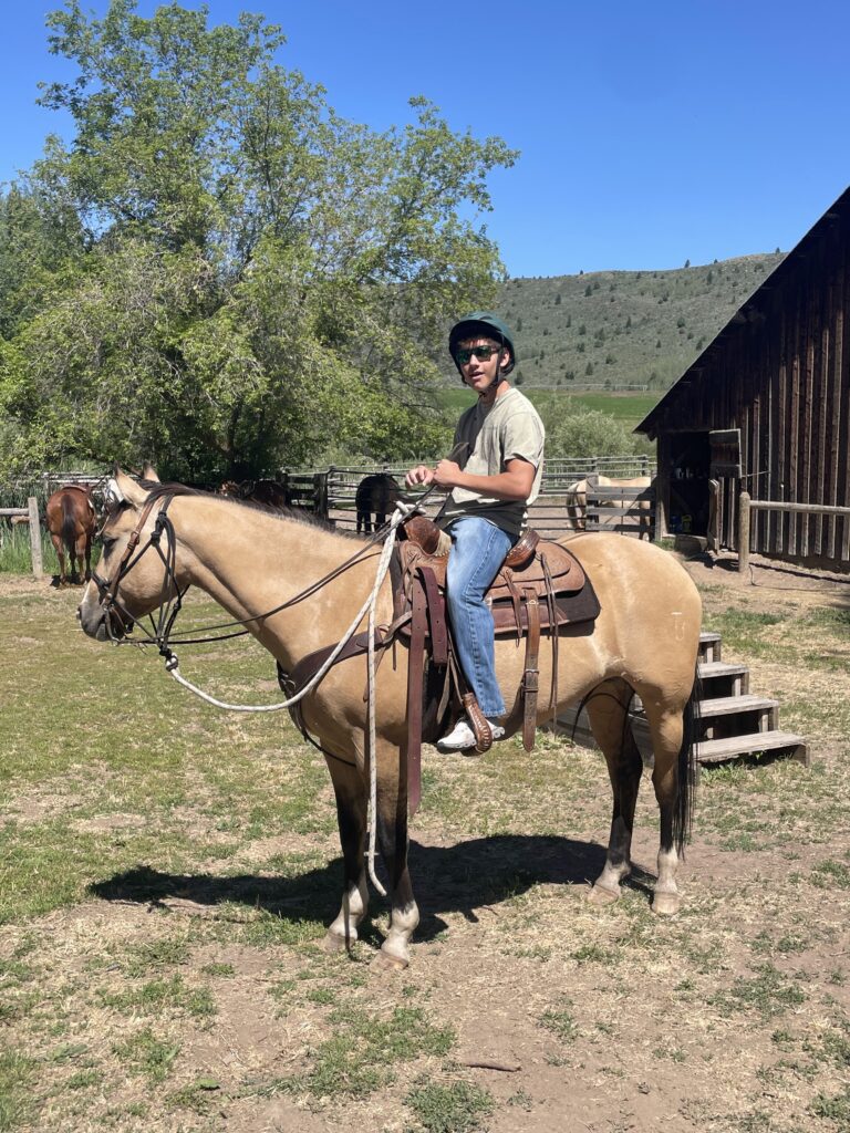 Student wearing a helmet and sunglasses sitting on a palomino horse near a ranch barn, with forested hills in the background