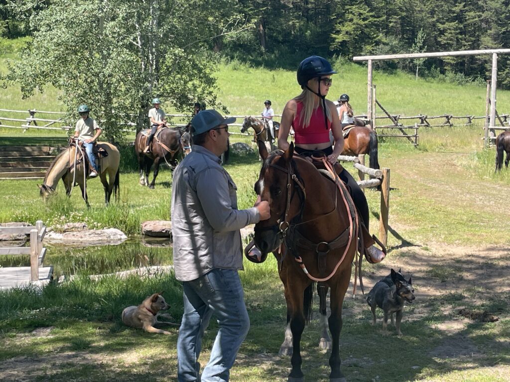 Ranch instructor guiding a student on a brown horse near the corral, with other riders visible in the background and ranch dogs resting nearby