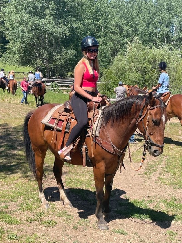 Student wearing a helmet and sunglasses confidently riding a chestnut horse, with other riders and horses visible in the background at the ranch