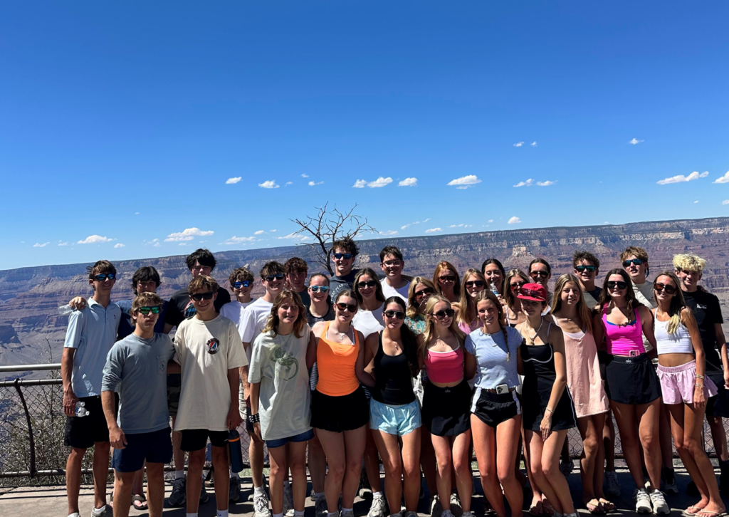 Large group of tour travelers standing together at a canyon overlook with a wide desert landscape and blue sky in the background.