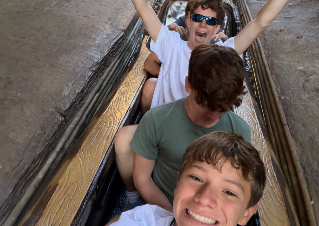 Three teenage boys cheering with arms raised while riding a log flume boat ride.