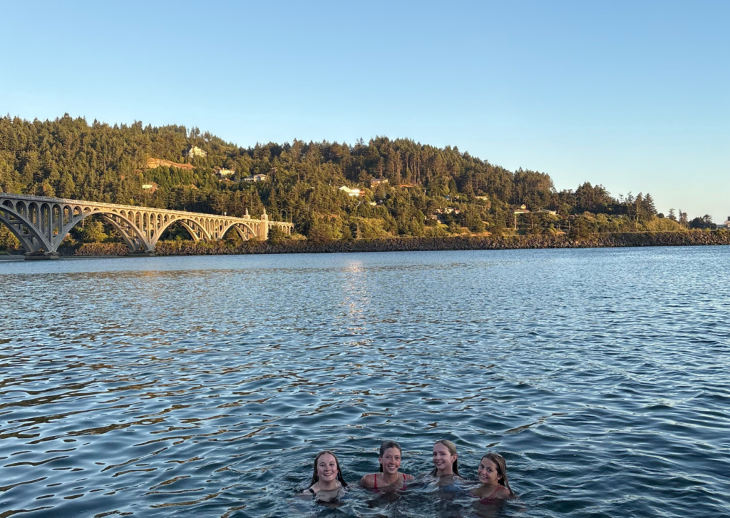 Group of women swimming in the Rogue River with an arched bridge and forested hills in the background