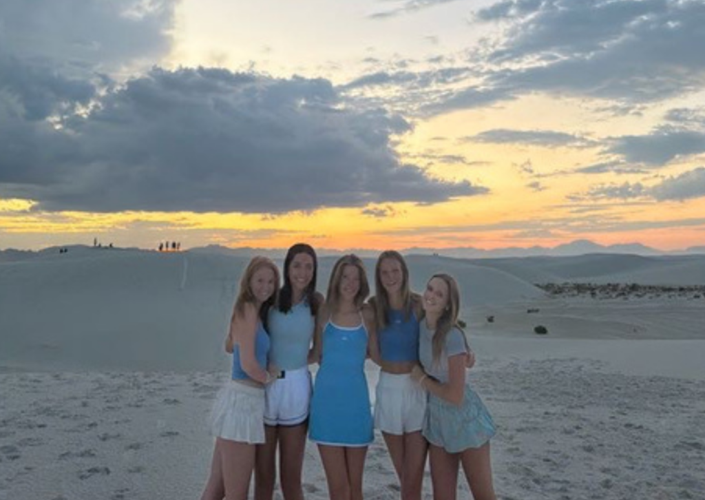 Five young women in matching blue and white outfits pose together on the dunes as a colorful sunset breaks through stormy clouds.