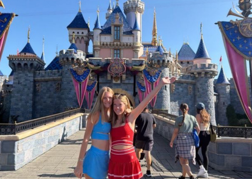 Two friends smiling and posing together in front of Sleeping Beauty Castle at Disneyland, one waving up at the castle.