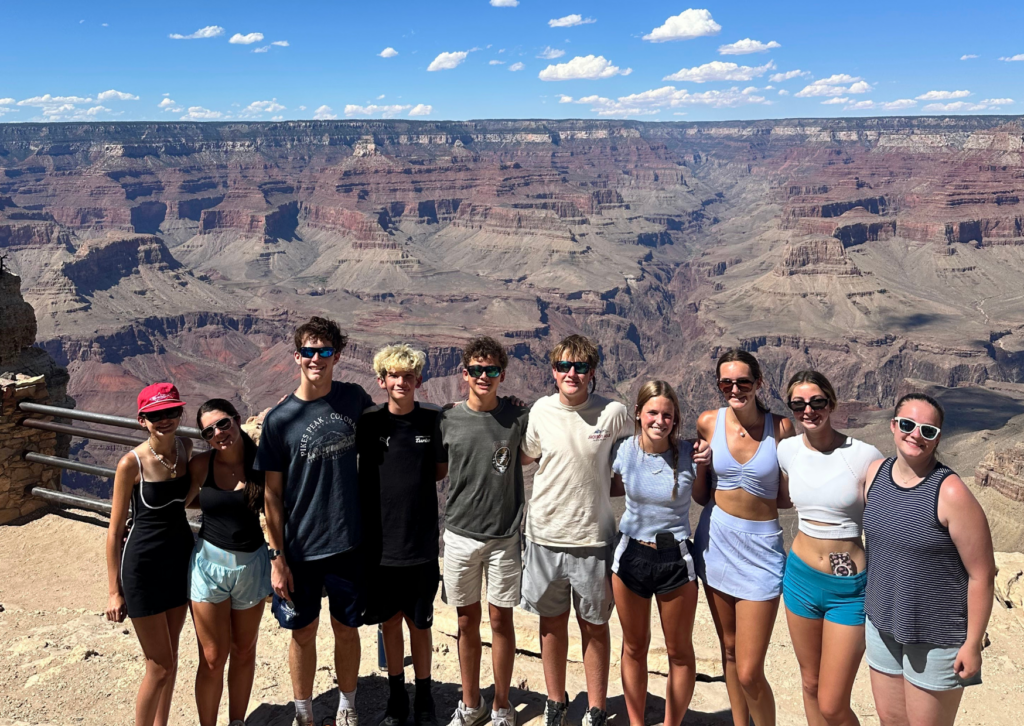 Group of tour travelers posing together at a scenic overlook of the Grand Canyon, with layered red rock canyon walls and a blue sky with clouds in the background.