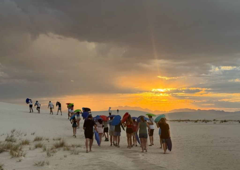 A line of people carrying colorful sleds trek across the white dunes toward a bright sunset breaking through stormy clouds.