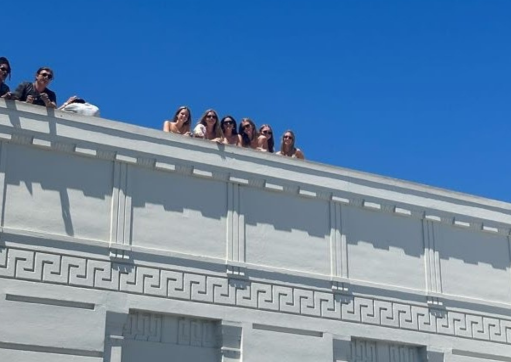 Group sitting along the rooftop ledge of a building with Greek key trim against a clear blue sky