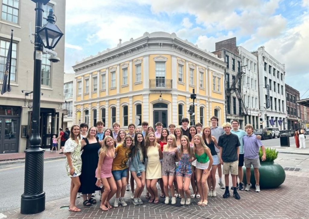 Group photo of tour participants standing on a street corner in the French Quarter of New Orleans, in front of a yellow historic building with wrought-iron balconies.