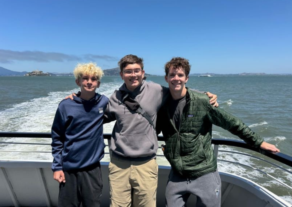 three boys looking at camera on ferry with water in background